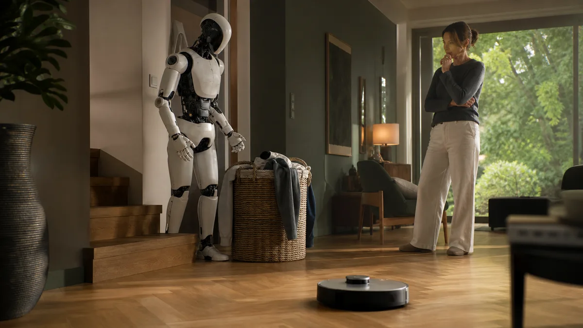 Propriétaire comparant un robot humanoïde encore limité par l’escalier à un robot aspirateur déjà opérationnel dans son salon.